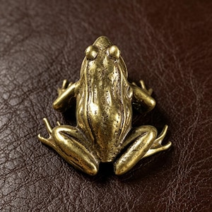 Può includere: Statuetta di rana in ottone dorato, con superficie strutturata. La rana è raffigurata seduta, con le zampe piegate e le braccia lungo i fianchi. Gli occhi della rana sono prominenti e il design è dettagliato.