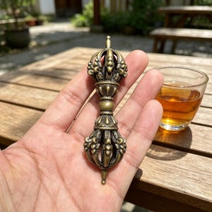 Peut inclure: Un Vajra de couleur bronze, un objet rituel, est tenu dans une main. Le Vajra présente des motifs floraux et géométriques ornés. Un verre de liquide ambré est posé à proximité sur une table en bois. L'arrière-plan montre un cadre extérieur flou.