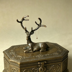 May include: A bronze-coloured deer figurine with antlers, resting on a decorative, antique-style box. A delicate necklace hangs from the antlers. The box has an ornate, detailed design and a clasp closure.