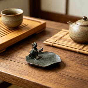 Könnte beinhalten: Eine bronzene Froschfigur auf einer blattförmigen Schale, die auf einem Holztisch steht. Eine Keramik-Teetasse und eine Teekanne sind ebenfalls zu sehen, was auf eine Teezeremonie hindeutet. Der Frosch hat ausgestreckte Arme.