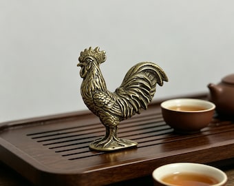 May include: A detailed brass rooster figurine stands on a wooden tea tray, with two small brown teacups filled with tea. The rooster is intricately designed, showcasing its comb, feathers, and stance. The tea set suggests a focus on tea ceremonies or decorative items.