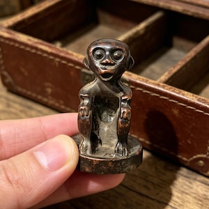 May include: A small, dark brown monkey figurine with a surprised expression. The figurine is held in a hand, with a brown leather box in the background. The monkey has a round base and detailed features.