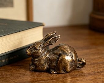 May include: A bronze-colored rabbit figurine in a resting pose. The sculpture has detailed features, including long ears and a curled tail. The rabbit is positioned on a wooden surface, suggesting a decorative item for a home or office.
