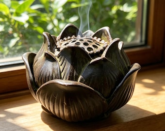 Peut inclure: Un brûleur d'encens en céramique brun foncé en forme de fleur de lotus en pleine floraison. Le centre présente un couvercle en dôme perforé avec un bord perlé, laissant s'échapper de fines volutes de fumée blanche depuis les pétales sculptés.