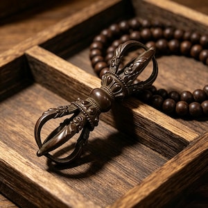 Könnte beinhalten: Ein bronzefarbener Vajra, ein rituelles Objekt, liegt in einer Holzschale. Eine Kette aus dunkelbraunen Gebetsperlen befindet sich im Hintergrund. Der Vajra hat komplizierte Details und ein symmetrisches Design. Die Holzschale hat eine natürliche Maserung.