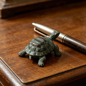 May include: A small, bronze-colored turtle figurine with detailed shell and limbs. The turtle is positioned on a wooden surface next to a pen. The figurine has a weathered appearance, suggesting an antique or vintage style.