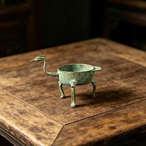 May include: A small, antique-style bronze vessel with a bird-like design. The vessel has a round bowl supported by three legs and a long neck and head resembling a bird. The bronze has a green patina.