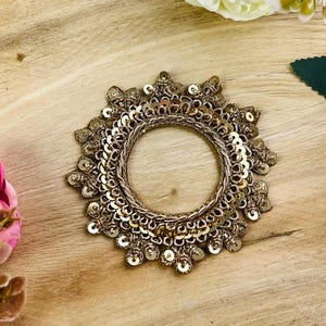 May include: A gold-coloured, circular decorative item with a beaded design. The item is embellished with small, round sequins and intricate detailing, set against a wooden surface. The overall aesthetic is ornate and elegant.