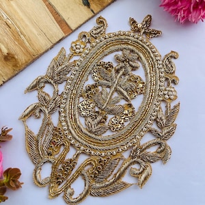 Peut inclure: Un écusson brodé doré de forme ovale avec un travail complexe de fils métalliques, de paillettes et de petites perles blanches. Le motif présente une fleur centrale entourée d'une bordure ornée de cordons dorés torsadés et de feuilles.