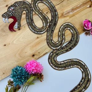Puede incluir: Una gran aplicación decorativa de serpiente elaborada con intrincadas cuentas y lentejuelas metálicas en tonos oro, bronce y plata. La serpiente se muestra en una postura sinuosa en forma de S con la boca abierta, colmillos blancos y lengua roja.