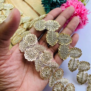May include: Gold-colored floral lace trim with intricate sequin detailing. The trim features a delicate floral and leaf design, perfect for embellishing garments or craft projects. The trim is held in a hand, with colorful flowers in the background.