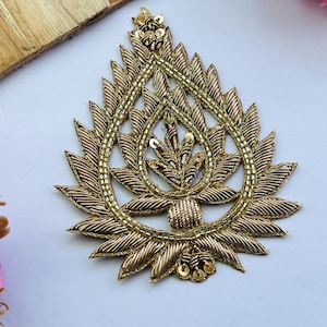 May include: Gold-colored embroidered applique in a teardrop shape. The applique features intricate detailing with gold thread, sequins, and a beaded border. The design has a central floral motif and a textured, layered appearance, suitable for embellishing garments or crafts.