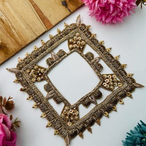 Könnte beinhalten: Ein quadratischer Zieraufnäher mit aufwendiger gold- und silberfarbener Metallstickerei, akzentuiert durch kleine kreisförmige Pailletten und Perlen. Das Design umfasst einen geometrischen Rand mit spitzen Kanten und detaillierten Fadenarbeiten.