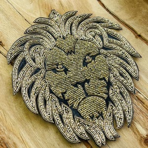 May include: Gold beaded lion head applique with a black backing. The lion's face is intricately detailed with gold beads, and the mane is composed of layered, curved gold beaded elements. The applique is set against a wooden background.