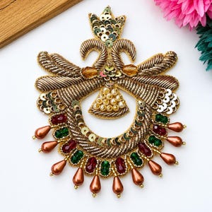 May include: A decorative embroidered patch featuring gold metallic thread, sequins, and beads. The design includes a central triangular cluster of cream pearls, surrounded by red and green faceted crystals and dangling copper-colored teardrop beads.