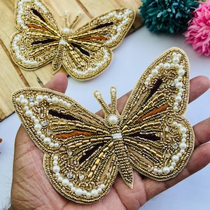 May include: Two butterfly-shaped appliques. The appliques are gold with intricate beadwork, including white pearls and gold beads. The wings have brown and orange accents. One applique is held in a hand, and the other is on a wooden surface.