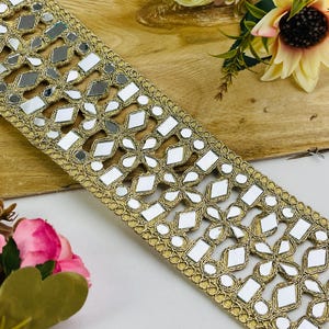 May include: Gold decorative trim with mirrored details. The trim has a repeating pattern of mirrored rectangles and diamond shapes, set within a gold border. This trim is suitable for embellishing clothing or craft projects.