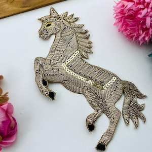 Puede incluir: Un parche con forma de caballo elaborado con hilo dorado metálico y lentejuelas. El diseño presenta un caballo encabritado con crines y cola detalladas, además de acentos negros en los cascos. El ojo está resaltado con hilo amarillo.
