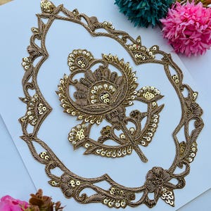 May include: A gold embroidered applique featuring a central floral motif surrounded by an ornate, pointed oval frame. The design is crafted with metallic gold thread and accented with small, reflective gold sequins throughout the pattern.