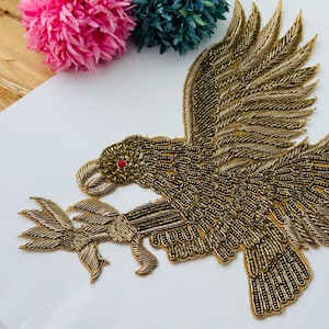 Puede incluir: Un parche con forma de águila hecho de hilo y cuentas de color oro metálico, con una pequeña cuenta roja para el ojo. El ave aparece en vuelo con las alas extendidas, sujetando una rama con sus garras, mediante un bordado detallado.