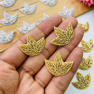 May include: A collection of small, leaf-shaped embroidered patches featuring metallic beadwork. The patches are available in silver and gold tones, each with a pointed, three-lobed design. The surface is covered in intricate, thin metallic beads.