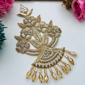 May include: A gold embroidered floral applique featuring metallic thread, sequins, and small white beads. The design includes five flower motifs with central crystals and a fan-shaped base adorned with dangling amber-colored teardrop beads.