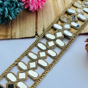 Puede incluir: Ribete decorativo dorado con detalles de espejo. El ribete presenta un patrón repetido de formas de espejo, incluyendo cuadrados y óvalos, dentro de un marco dorado. El ribete está sobre una superficie clara, con flores decorativas rosas y turquesas en el fondo.