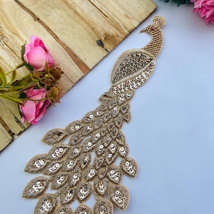 Puede incluir: Una aplicación de pavo real en tono dorado con bordados intrincados y detalles en hilo metálico. El diseño está adornado con numerosas lentejuelas plateadas reflectantes dispuestas en un patrón de abanico sobre las plumas y el cuerpo.