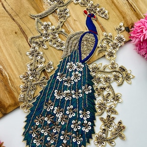 Peut inclure: Une application décorative en forme de paon avec une broderie en fils bleu profond et sarcelle, rehaussée de paillettes métalliques dorées et de perles. Le motif représente un paon entouré de branches florales ornées de fleurs à paillettes dorées.