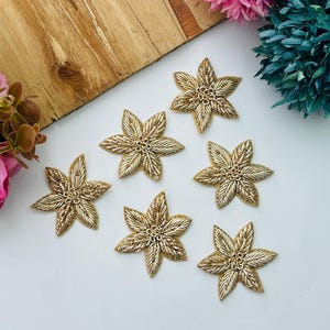 以下が含まれることがあります： 質感のあるメタリック糸のディテールが施された、6つの金色の星型フラワーアップリケ。各ピースは6枚の尖った花びらと中央の円形要素で構成されており、滑らかな刺繍とビーズ状の金色の刺繍が組み合わされています。