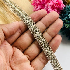 May include: A metallic gold decorative trim featuring a complex braided pattern with a textured, woven appearance. The trim has a consistent width and is finished with small, looped edges along both sides, creating a detailed border design.