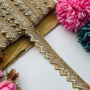 May include: A gold metallic trim featuring a scalloped edge adorned with small, reflective sequins. The decorative ribbon displays intricate embroidery and a repeating zigzag pattern, showcasing a shimmering finish throughout the entire length.