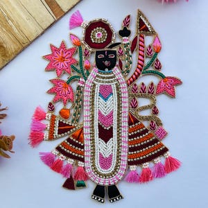 May include: An ornate, embroidered applique featuring a figure with a black face and elaborate attire. The design incorporates pink, white, and gold beads, with accents of red, green, and orange. Pink tassels and floral elements complete the detailed, handcrafted look.
