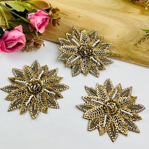May include: Three gold-colored, flower-shaped embellishments with intricate sequin and bead detailing. The floral designs are arranged on a light-colored wooden surface, with pink and white flowers in the background.