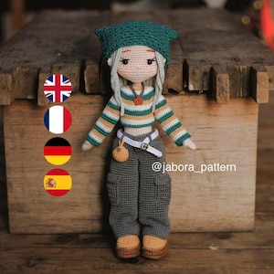 Puede incluir: Muñeca de ganchillo hecha a mano con gorro verde, camisa a rayas y pantalón gris. La muñeca tiene pelo gris y una pequeña bolsa. La muñeca está de pie frente a una caja de madera. La muñeca mide unos 25 cm de alto.