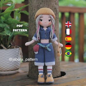 May include: A crochet doll wearing a tan beanie, a blue collared crop top, and matching blue shorts with a light blue belt. The doll has long hair with pink highlights, white socks with red and blue stripes, and tan shoes. Text reads PDF PATTERN.