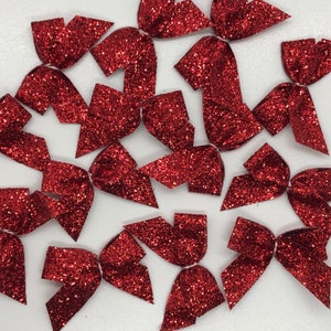 May include: A collection of small, red glitter bows scattered on a white surface. The bows are cut into various shapes, each with a sparkling, textured finish. The bows are likely for crafting or embellishment.