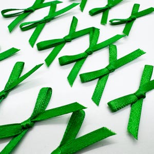 May include: A collection of small, bright green ribbon bows made from a smooth, woven fabric. Each bow features two loops and two short, angled tails, arranged in a scattered pattern across the frame, showcasing a consistent texture and color.