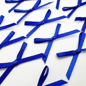 May include: A collection of small, vibrant royal blue ribbon bows made from smooth satin material. Each bow features a central knot with two short, angled tails extending outwards, arranged in a scattered pattern across the frame.
