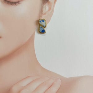 Puede incluir: Un pendiente de botón que presenta dos piezas de cerámica blanca de formas orgánicas e irregulares con motivos florales azules. Cada fragmento de cerámica está enmarcado por un fino borde metálico dorado, creando un diseño asimétrico.