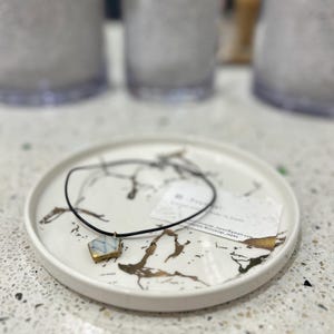以下が含まれることがあります： 白と青のひび割れ模様に金色の縁取りが施された五角形のペンダントで、細い黒いコードに吊り下げられています。ペンダントは、黒と金のマーブル模様の筋が入った白い丸皿の上に置かれており、小さな白いカードも添えられています。