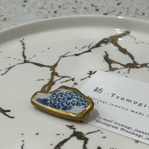 May include: A small, irregular ceramic fragment featuring a blue and white patterned surface with gold-colored edges, displayed next to a white card with the text 紡 - Tsumugi - kintsugi jewelry made in Japan, e-mail tsumugi_japan, Instagram @tsumugi_japan.