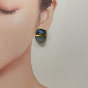 May include: A stud earring featuring two stacked, irregular shapes encased in a gold-toned metal border. The top section displays a white and blue vertical striped pattern, while the bottom section is a smooth, solid light blue, matte-finish piece.