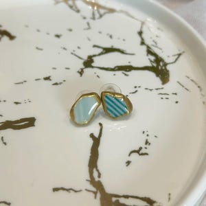 以下が含まれることがあります： 金色の縁取りが施された不規則で滑らかな形状のスタッドピアス一対。片方は無地のライトブルーで、もう片方はライトブルーと白のストライプ模様。どちらのピアスにも透明なプラスチック製のキャッチが付いています。