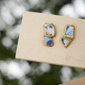 May include: A pair of stud earrings featuring irregular shards of white ceramic with blue floral patterns, each piece edged in gold-toned metal. The earrings are displayed on a light tan card with the text Tsumugi printed in the bottom right corner.