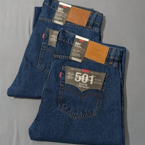 May include: Three pairs of dark blue denim jeans with tags attached. Each pair has a brown leather patch and a red tag on the back pocket. The jeans are folded neatly, showcasing the back pockets and tags.