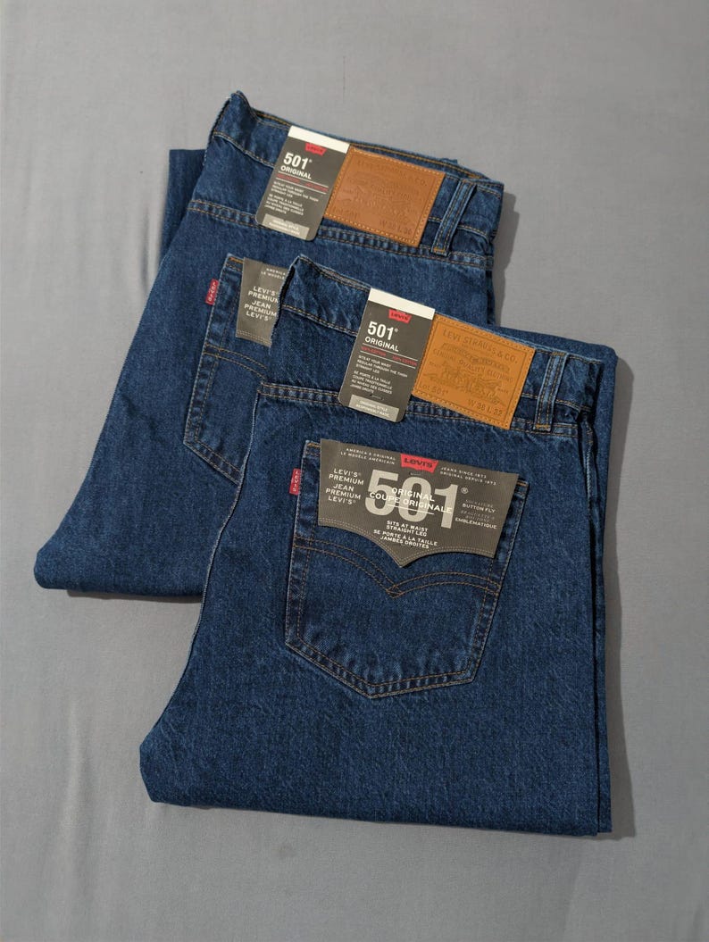 May include: Three pairs of dark blue denim jeans with tags attached. Each pair has a brown leather patch and a red tag on the back pocket. The jeans are folded neatly, showcasing the back pockets and tags.
