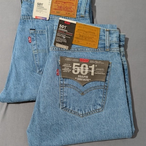 May include: Two pairs of light blue Levi's 501 Original jeans. Each pair has a classic five-pocket design, a red Levi's tag on the back pocket, and a leather patch. The jeans have tags with the Levi's logo and size information. One pair is W34 L30, the other is W34 L32.