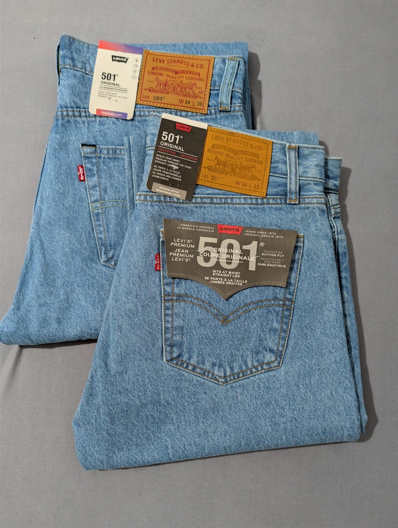 May include: Two pairs of light blue Levi's 501 Original jeans. Each pair has a classic five-pocket design, a red Levi's tag on the back pocket, and a leather patch. The jeans have tags with the Levi's logo and size information. One pair is W34 L30, the other is W34 L32.