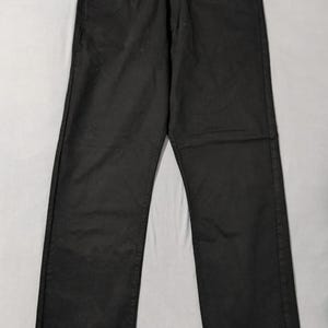 May include: Black denim jeans with a classic five-pocket design. The jeans have a button and zipper closure, belt loops, and copper rivets. The garment is laid flat on a light gray surface.